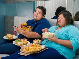 a photo of two obese people eating in front of tv