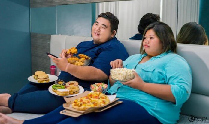 a photo of two obese people eating in front of tv